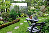 View past weathered wooden table and chair to greenhouse and flowerbeds in summer garden