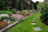 Blühende Blumen im Beet mit Steineinfassung und Trittplatten in gemähter Rasenfläche im Garten