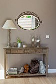 Table lamp with white lampshade and candle lanterns on rustic wooden console table below oval mirror on wall painted pale grey