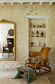 Wicker chair and footstool in front of glass shelves in niche in stone wall next to gilt-framed mirror