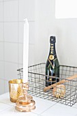White candles in gilt candlestick and gold tealight holders in front of bottle of Champagne in wire basket