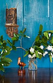 Carved pelican figurine and flowering branches in vase below wooden ethnic carvings on wall painted light blue