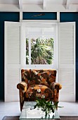 Armchair with brown, retro floral pattern and wicker table with glass top in front of French window with interior folding shutters