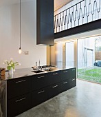 Designer kitchen counter with black drawers and stainless steel handles below extractor hood in minimalist interior; view through floor-to-ceiling window in background