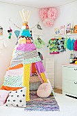 Teepee made from colourful and patterned fabric remnants in front of child's clothing and flower decorations hung on wall in corner