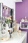 Schminkutensilien auf weißem Wandtisch vor Spiegel mit seitlich angeordneten Leuchten, im Hintergrund violett getönte Wand