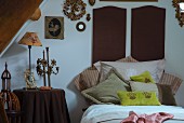 Drapierte Kissen vor dunkelbraunen Bettkopfteilen an Wand, seitlich nostalgische Kerzenhalter und Tischleuchte auf Beistelltisch
