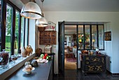 Grey kitchen worksurface below bank of windows and vintage cooker below interior window with view into rustic living area