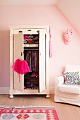 Child's clothing in wardrobe with open doors next to white armchair against pink-painted wall