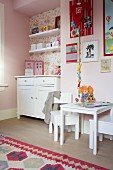 Child's bedroom with white furnishings, table and chairs against pink wall, chest of drawers and shelves in wallpapered niche