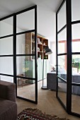 Stahl-Glaswand mit offener Tür und Blick in Wohnbereich auf Schrank und Stehleuchte neben Sofa