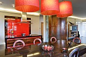 Orange lamps above metal chairs and large table with dark, exotic wood veneer; open-plan kitchen with red fitted cupboards in background