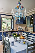 Dining table, pale grey chairs and chandelier made from coloured glass in kitchen with cupboards painted blue