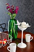 Still-life arrangement of green glass vase, retro wire basket, white china mugs with knitted surface texture and candlestick