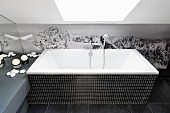 Badewanne mit schwarzen Mosaikfliesen vor Fototapete mit Bergmotiv, darüber Dachflächenfenster