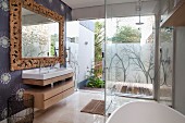 Glazed, floor-level shower opposite washstand and mirror with solid wooden frame carved with leaves in modern bathroom; view onto terrace through glass wall