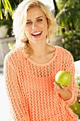 A blonde woman wearing an apricot, open-work jumper holding a green apple