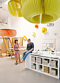 Verschiedene Pendelleuchten mit grossem Schirm aus farbigen Lamellen, im Hintergrund Frau und Mann an Arbeitstischen, in der Werkstatt