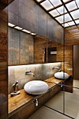 Elegant, contemporary bathroom with countertop basin on wooden counter in niche with antique-brass-effect tiles on wall and indirect lighting