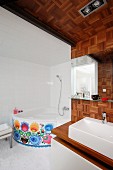 Modernes Bad, teilweise mit Mosaik Holzplatten an Wand und Decke ausgelegt, eingebauter Waschtisch, im Hintergrund Eckbadewanne mit dekorativem Blumenmuster an Frontseite