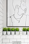 Modern line drawing portrait on tiled wall in bathroom above row of decorative grasses in zinc pots on masonry shelf