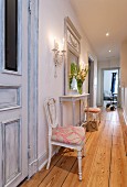 Long narrow hallway with rustic wooden floor; two antique re-upholstered chairs flanking delicate white console table below mirror