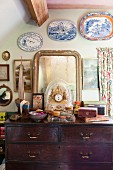 Table clock and vintage mirror on top of wooden chest of drawers below decorative plates on wall