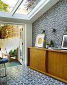 Fifties-style, pale wooden sideboard against patterned wallpaper and patterned tiled floor in front of terrace doors