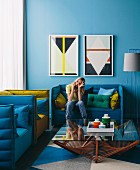Woman on blue sofa behind glass table and framed pictures with geometric patterns on blue wall