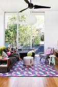 Corner sofa and retro armchairs on colourful rug with graphic pattern in front of floor-to-ceiling windows overlooking garden