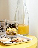 Orangensaft in Trinkgläsern mit grafischem Motiv & dazu passendem Tablett auf gelbem Nachttischchen
