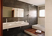Blick ins Designerbad, weisser Waschtisch mit zwei Schüsseln vor dunkel gefliester Wand, seitlich Podest mit Holzboden vor Badewanne