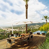 Dining set and parasol on wooden deck with view of town on hillside; infinity pool in background