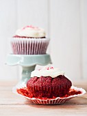 Two Red Velvet cupcakes with white frosting on a plate and on a cake stand