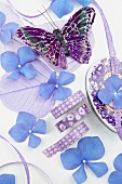 Blau-lilafarbenes Stillleben mit Hortensienblüten, Dekoschmetterling, Dekoblättern & Schleifenbändern