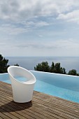 Designer chair made of white plastic on wooden deck next to infinity pool with sea view