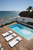 Pale futon sun loungers with matching pillows on wooden deck surrounding pool with ocean in background