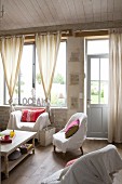 Various armchairs with white loose covers around coffee tables in front of large window with knotted curtains in simple living room