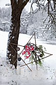 Dekostern aus Holzstäben am Baum lehnend, mit Weihnachtskugeln und Schleife geschmückt, davor brennende Kerzen im Schnee