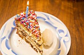 A colourful slice of birthday cake with a burning candle and a scoop of vanilla ice cream