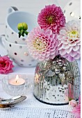 Pink dahlias in mercury glass vase on lace doily with teacups in background