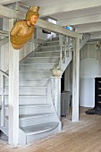 Old ship's figurehead hung on wooden structure in front of winding wooden staircase painted pale grey in rustic interior