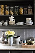 Vase of flowers on kitchen counter below white crockery and storage jars in open-fronted wall cabinets