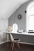 Classic chair with white shell seat at desk below window in grey-painted wall of attic room with arched ceiling