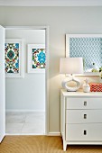 Retro table lamp on white chest of drawers next to open door showing view of framed ornamental patterns