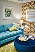 Cups on tray on blue, velvet, round ottoman and pale blue sofa in living room; ornately patterned wallpaper on accent wall