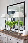 Elegant silver table lamps with white lampshades on grey apothecary cabinet below framed mirror in elegant interior