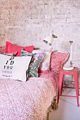 Bett mit rotweiß gemusterter Tagesdecke und Kissen, seitlich rosa lackierter Klassiker-Metallhocker im Retrostil mit Nachttischlampe vor Ziegelwand