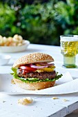 A hamburger and a sesame seed bun with lettuce, mustard, ketchup, tomatoes and red onions on a picnic table with beer and chips
