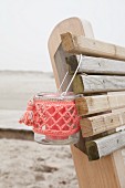 Candle lantern with coral-pink crocheted cover hung from backrest of wooden bench on beach